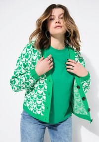 Cardigan verde con motivo a cuori bianchi, chiusura a bottoni, polsini a coste e una t-shirt verde uniforme sotto. Indossato con jeans blu.