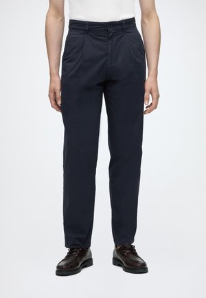 Personne portant un pantalon droit bleu marine foncé avec plis, des chaussures en cuir noires à lacets et un haut blanc sur un fond uni.