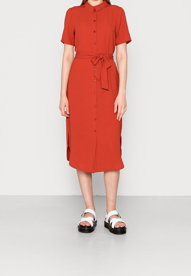 Abito camicia rosso a maniche corte con apertura a bottoni e fessure laterali, cinte in vita da una cintura in tessuto. Indossato con sandali bianchi.