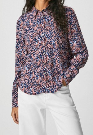 Vrouw draagt een marineblauw overhemd met knopen en een kleine roze bloemenprint, en witte broek, met één hand in de zak, tegen een effen achtergrond.