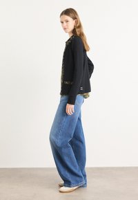 Zwarte cardigan met een gepatineerde kraag en manchetten, gecombineerd met losse blauwe spijkerbroek en witte sneakers op een lichte achtergrond.
