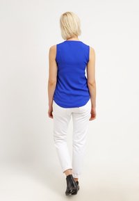 Sininen hihaton toppi, joka on valmistettu sileästä kankaasta, yhdistettynä valkoisiin housuihin. Asu on istuva ja siinä on hienovarainen kaareva helma. Mustat nilkkurit.