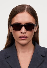 Lunettes de soleil noires rectangulaires avec une finition brillante et des branches épaisses en écaille. Portées par un mannequin dans une veste de costume anthracite.