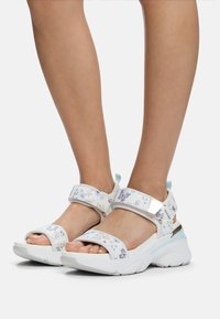 Hvide sandaler med blomsterprint, der har justerbare stropper, polstrede såler og en kraftig hæl. Har metalaccenter og en struktureret overflade.