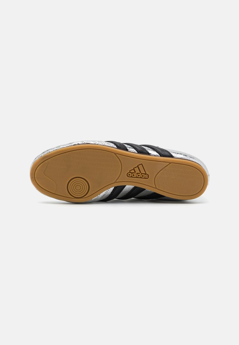 Suela de zapatilla deportiva negra y plateada con tres rayas negras, un patrón texturizado y un diseño de banda circular con un logo.