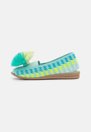 Zapatos planos tejidos y coloridos con un patrón de zigzag en turquesa, amarillo y verde, con una suela marrón texturizada y un pompón decorativo.