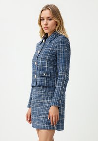 Kobieta w niebiesko-białej koszulowej kurtce w kratę z złotymi guzikami i dopasowanej spódnicy o wysokim stanie, stojąca na jednolitym tle.