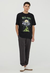T-shirt nera in cotone con grafica "Rick and Morty" e design di UFO. Abbinata a pantaloni della tuta grigi e sneakers bianche con suola marrone.
