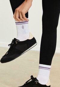 Persona che indossa leggings neri e calzini bianchi a coste con strisce viola e un piccolo logo, mentre aggiusta un calzino sopra le sneakers nere.