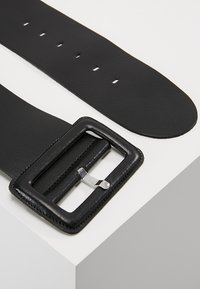 Cintura in pelle nera con fibbia rettangolare in argento. Presenta diversi fori per le regolazioni, una texture liscia e cuciture a contrasto sui bordi.