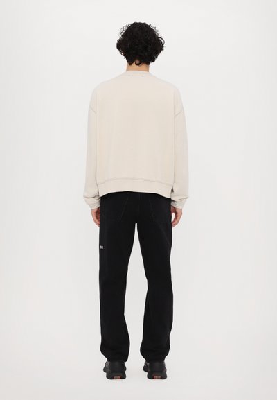 Sweatshirt beige clair avec des poignets et unourlet côtelés, associé à un pantalon noir arborant un logo discret à l'arrière. Tissu texturé, coupe décontractée.