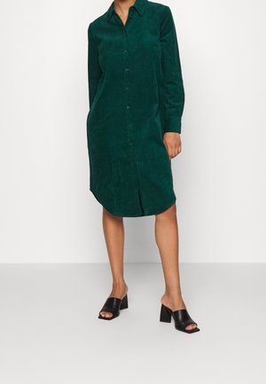 Žena stojící na bílé podlaze, oblečená v dlouhých rukávech tmavě zelených manšestrových šatech na knoflíky a černých nazouvacích botách na blokovém podpatku s otevřenou špičkou.