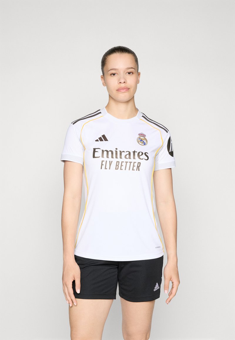 Biela futbalová dres s čiernymi a žltými akcentmi, s emblémom Real Madrid a logom "Emirates Fly Better". Nosený s čiernymi šortkami.