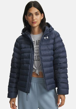 Frau trägt eine marineblaue gesteppte Daunenjacke mit Kapuze, graues geknotetes T-Shirt und hellblaue Jogginghose, schaut nach vorne mit neutralem Gesichtsausdruck.