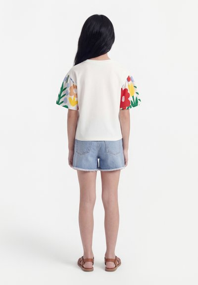 Κορίτσι στέκεται με την πλάτη γυρισμένη, φοράει λευκό πουκάμισο με πολύχρωμα φλοράλ μανίκια, ανοιχτό μπλε τζιν σορτς και καφέ σανδάλια.