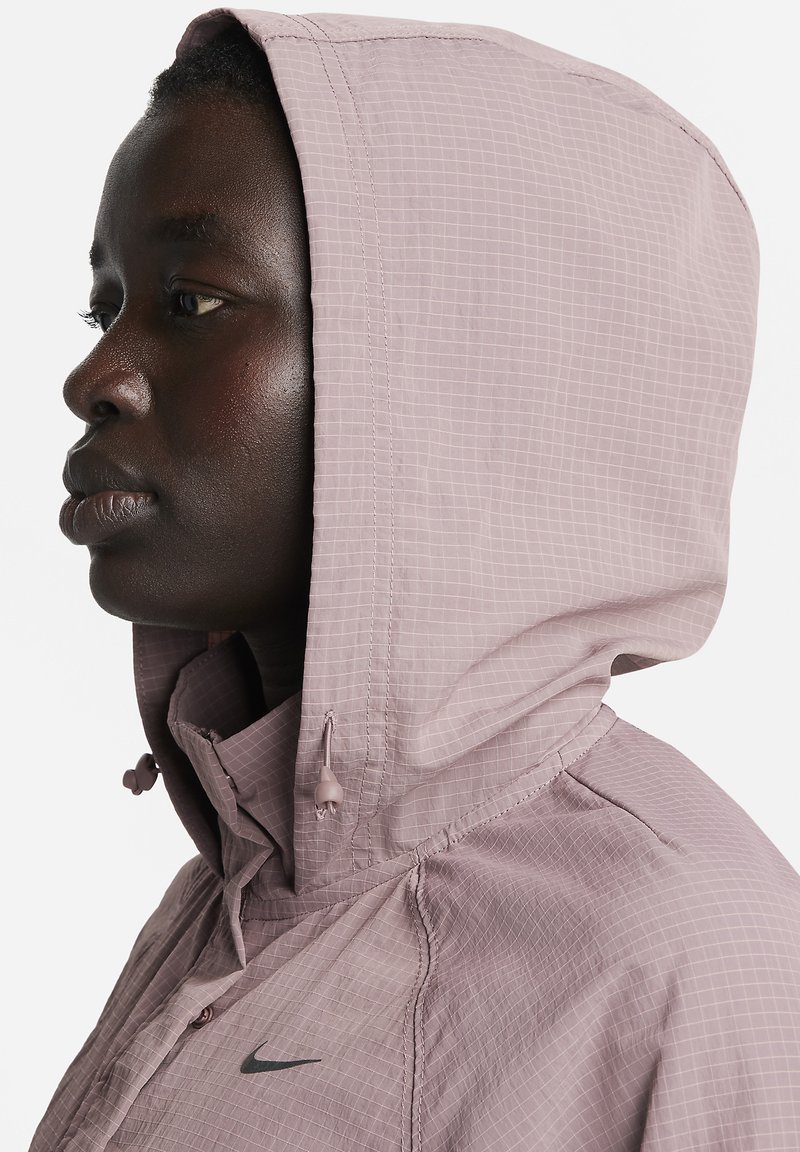 Lichtroze hooded jack met een ruitpatroon, verstelbare trekkoorden en het Nike-logo op de linkerborst. Lichtgewicht stof, close-up weergave.
