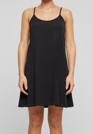 Woman wearing a sleeveless black A-line dress with thin straps, standing against a plain light gray background.