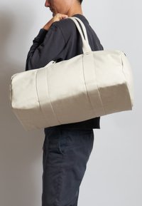 Beige canvas sporttas met dubbele handgrepen en twee horizontale banden. Ronde vorm, stevige textuur en ruime binnenkant voor opslag.