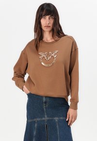 Bruine oversized sweatshirt met een ronde halslijn en een versierd grafisch ontwerp op de voorkant. Draagt met een donkere spijkerrok.