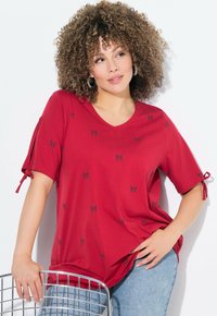 Haut rouge à manches courtes avec un col en V et des motifs de nœuds noirs. Présente des manches nouées, un tissu doux et une coupe décontractée. Porté avec un jean bleu.