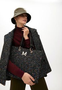 Kabelka z tvídu s černým a multikolorovým tkaním, zlatým kováním a černými uzlovými rukojeťmi; nošena s vínovým rolákem a vzorovaným kabátem.