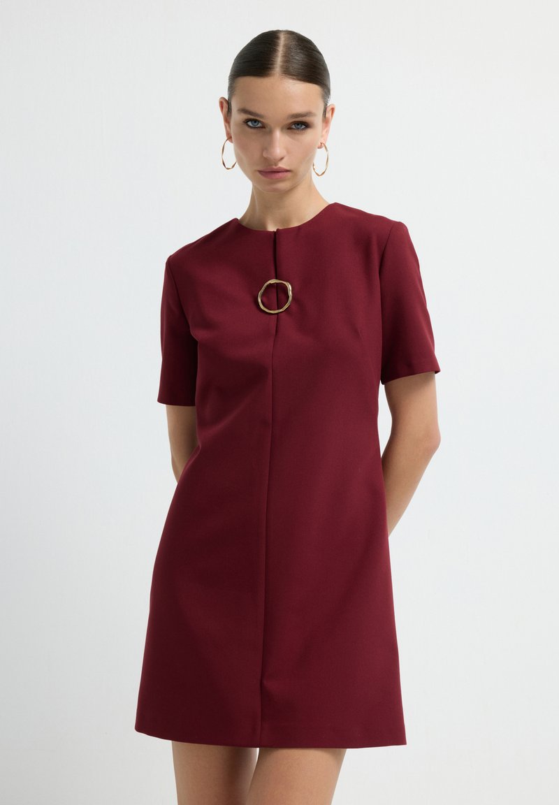 Robe bordeaux à manches courtes avec un col rond et un accent circulaire doré au niveau de la poitrine. Fabriquée en tissu lisse, à coupe droite.