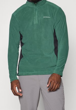 Mann mit grünem Columbia Fleecepullover mit Viertelreißverschluss und grauer Hose, stehend vor einfarbig hellem Hintergrund.