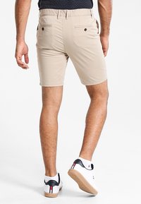 Beżowe bawełniane szorty o gładkiej fakturze, z bocznymi kieszeniami i dwiema tylnymi kieszeniami. Noszone z białymi sneakersami z czarnymi akcentami i beżową podeszwą.