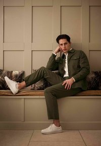 Oliven grøn jakke og bukser lavet af tekstureret stof, parret med hvide sneakers. Model siddende på en træbænk mod en beige væg.