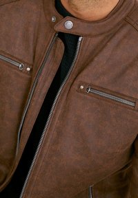 Giubbotto da moto in similpelle marrone con colletto alto, due tasche sul petto con zip e dettagli in metallo argentato.