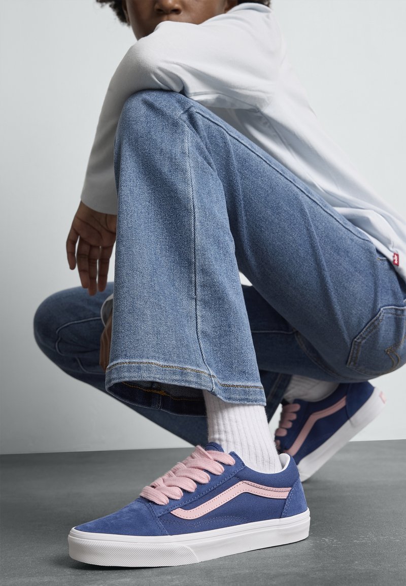 Personne accroupie portant un jean bleu en denim, des chaussettes blanches et des baskets bleu marine avec des lacets roses et des bandes latérales sur un sol gris.