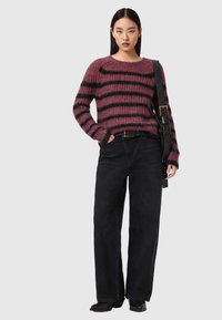 Femme portant un pull pelucheux violet et noir à rayures, un jean large noir, des bottes noires et un sac noir avec une large bandoulière portée sur une épaule.