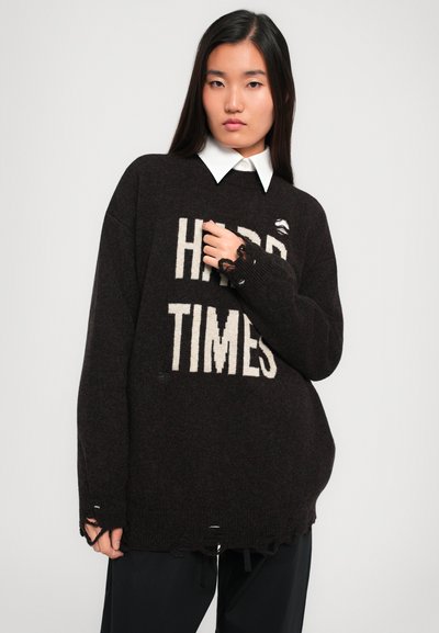 Oversized dark brown sweater with "HARD TIMES" printed in cream. Features distressed edges and detailing on cuffs. Layered over a white collar shirt.