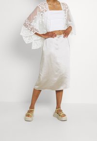 Kvinde iført en hvid blondes transparent cardigan over en hvid crop top, en cremet satin midi-nederdel og platform sandaler med gule remme.