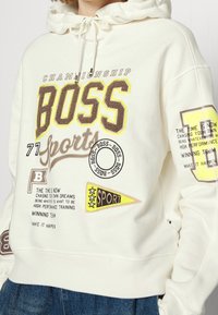 Sweat à capuche blanc avec texte et graphismes brodés en marron et jaune. Comprend une capuche avec cordon de serrage et plusieurs écussons, dont des motifs liés au sport.