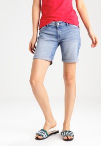 Pantalones cortos de mezclilla azul claro con dobladillos enrollados, que presentan un clásico diseño de cinco bolsillos. Combinados con sandalias deslizantes con estampado en verde y negro.