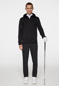 Svart zip-up huva jacka kombinerad med svarta byxor, har en slim fit och slät textur. Modellen håller en golfklubba och bär vita skor.