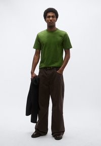 Πράσινο κοντομάνικο μπλουζάκι συνδυασμένο με άνετο καφέ παντελόνι και σκούρα παπούτσια. Το μοντέλο κρατάει ένα μαύρο σακάκι πάνω σε ένα μπράτσο.