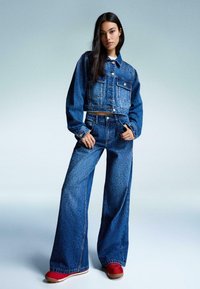 Jeans jakna i široke traperice u srednje plavoj s diskretnim ukrasima. Model nosi crvene tenisice. Glatka tekstura; skraćeni dizajn jakne.