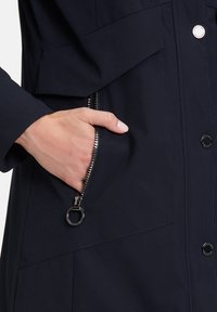 Veste bleu marine avec une poche zippée et des boutons pression. Le tissu est lisse avec une finition mate et des coutures minimales.