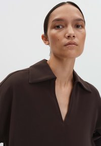 Femme aux cheveux noirs tirés en arrière, portant un t-shirt marron foncé à col en V large, avec une expression neutre, sur un fond clair.