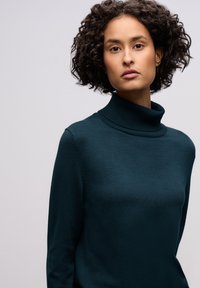 Tumma sinivihreä pooloneule, jossa on pehmeä pinta ja istuva siluetti. Siinä on korkea kaulus ja pitkät hihat, näkyy vähän saumoja.