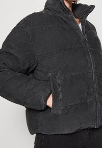 Svart kordfluwelpuffjacka med hög krage, horisontellt quiltad design, sidofickor och dragkedja. Mjuk textur med ruggad yta.