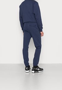Man med marinblå joggers och sweatshirt med svarta och vita Nike Air sneakers, stående med ryggen mot kameran framför en färgglad vit bakgrund.