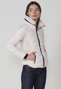 Padded bunda svetlej smotanovej farby s vysokým golierom, čiernym zipsom a bočnými vreckami. Obsahuje štruktúrované bočné panely a čierne akcenty.