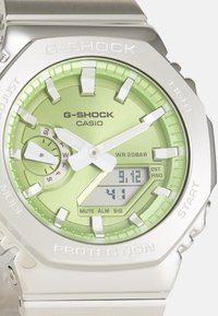 Relógio G-SHOCK com caixa em aço inoxidável, mostrador verde, detalhes em prata, mostrador analógico e digital, e rótulos de botões proeminentes. À prova d'água até 20 bar.