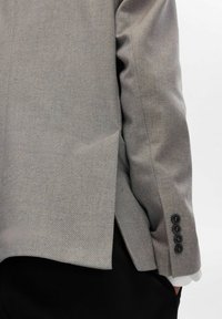 Grå herringbone-blazer detalj med en slits bak, skräddarsydd passform och fyra mörka knappar på ärmen. Texturerad tyg ökar djupet.