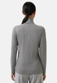 Kobieta o ciemnych włosach związanych w kucyk, ubrana w dopasowaną, szarą bluzkę z długim rękawem oraz jasnoszare spodnie, widziana od tyłu na jednolitym tle.