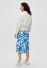 Lichtbeige bomberjack met een cropped ontwerp, gecombineerd met een middenlange blauwe rok met witte abstracte golvende patronen. Witte sneakers.