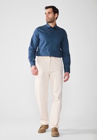 Pantalones beige claros, de corte holgado, combinados con una camisa de botones azul oscuro. El conjunto presenta un cuello con solapa y un discreto detalle de bolsillo.